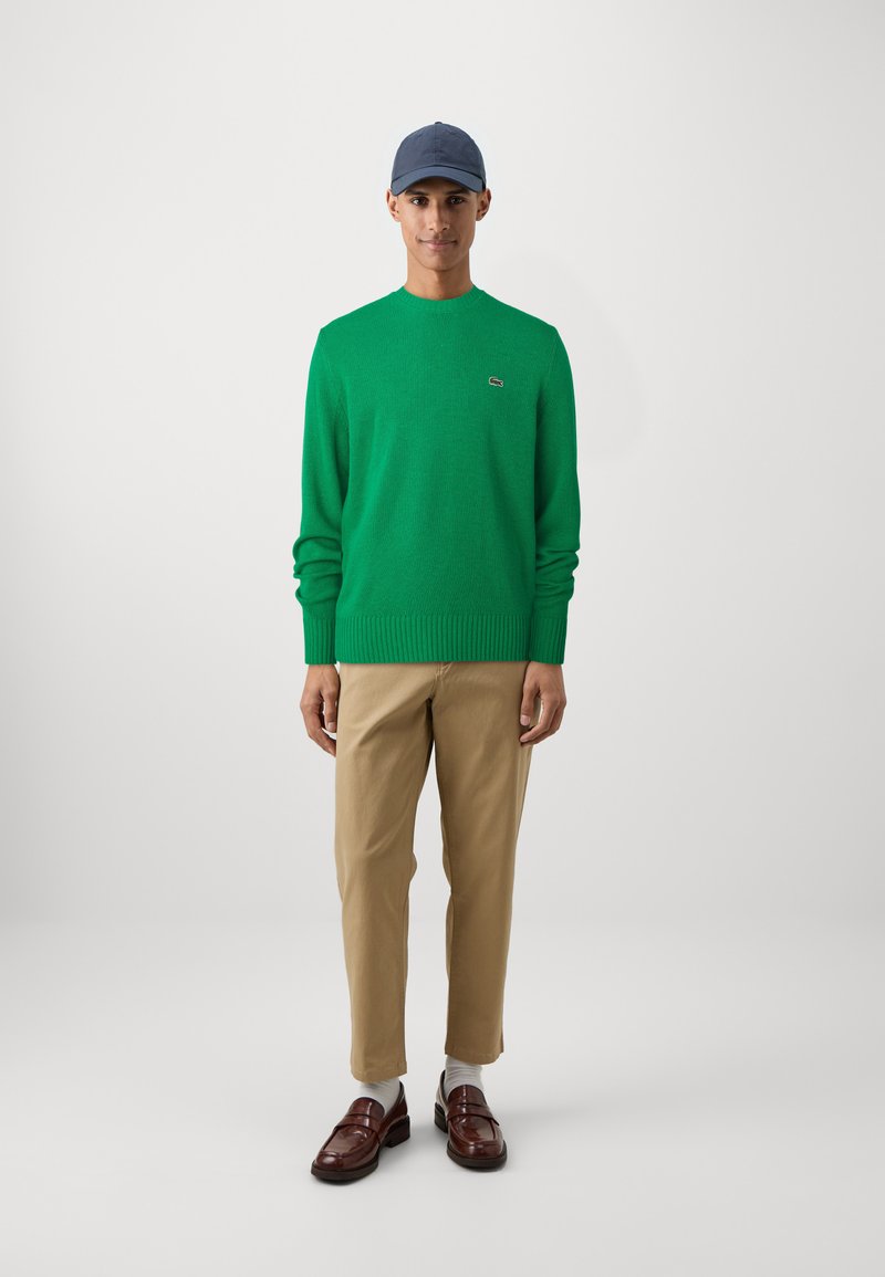 Maglione verde lavorato a maglia con polsini a coste, abbinato a pantaloni beige e mocassini marroni. Accessorio include un berretto blu navy.