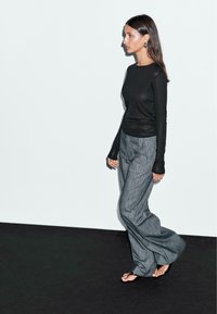 Long-sleeve black top with fitted silhouette and sheer texture, paired with gray wide-leg trousers featuring vertical stripes.