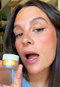 En klar burk med läppbalsam med ett vitt lock och en gul etikett, hålls framme av en person. Mikrokonsistens, krämig utseende, subtil glans.