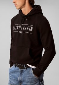 Άνδρας που φοράει μαύρο φούτερ Calvin Klein με λευκό λογότυπο και μπλε τζιν, με τα χέρια στις τσέπες, κοιτάζοντας στο πλάι με γκρι φόντο.