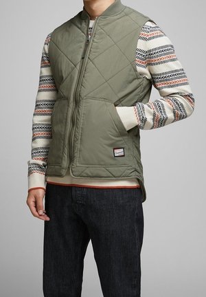 Mann trägt olivgrüne gesteppte Weste über einem weißen gemusterten Pullover mit schwarzen Jeans, Hand in der Westentasche, vor einem einfarbigen grauen Hintergrund.