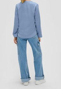 Maglia a maniche lunghe blu chiaro con orlo arrotondato, abbinata a jeans oversize blu chiaro e sneakers bianche, vista da dietro.