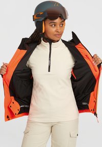 Femme portant un casque de ski noir et orange et une veste de ski orange, ouvrant sa veste pour révéler un pull à zip crème et un pantalon beige.