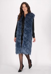 Lång, blå fuskpälsväst med en texturerad yta, bred krage och vertikala randmönster, bärs över en svart outfit och kombineras med klackar.