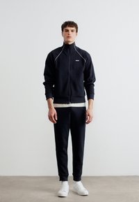 Νέος άνδρας που στέκεται φορώντας μαύρο μπουφάν με φερμουάρ, μαύρο παντελόνι, λευκή εσώρουχο και λευκά αθλητικά παπούτσια μπροστά σε έναν απλό λευκό τοίχο.