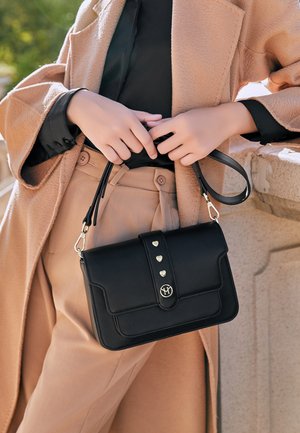 Femme portant un manteau beige et un pantalon tient un sac à main noir orné de clous en forme de cœur dorés et d'une fermeture circulaire avec logo.