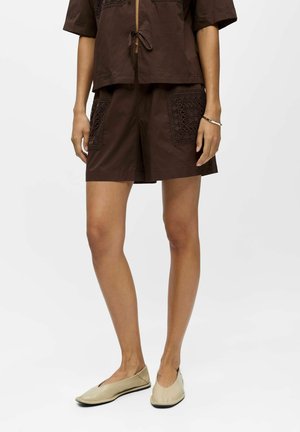 Femme portant un short marron foncé et un top assorti avec des poches brodées, des chaussures plates beiges et un bracelet en argent au poignet gauche.