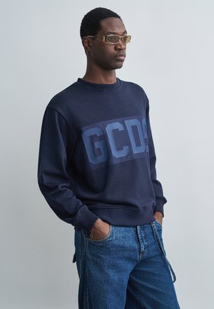 Felpa blu navy con un grande logo "GCDS" in una tonalità di blu leggermente più chiara. Tessuto morbido, scollatura a girocollo, polsini a costine, abbinata a jeans in denim.