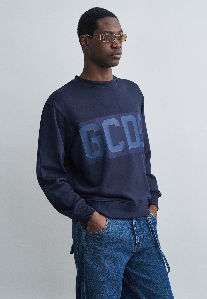 Marinblå sweatshirt med en stor "GCDS"-logotyp i en något ljusare blå nyans. Mjuk tyg, rundhalsad krage, ribbade ärmslut, tillsammans med denimjeans.