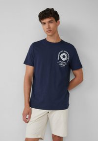 Himmelblaues Baumwoll-T-Shirt mit weißem Text und floralem Design auf der linken Brust. Rundhalsausschnitt, kurze Ärmel, kombiniert mit beigen Shorts.