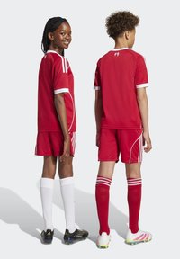 Två unga fotbollsspelare i röda och vita uniformer stående rygg mot rygg, en med ett leende som ser åt sidan, den andra vänd bort.