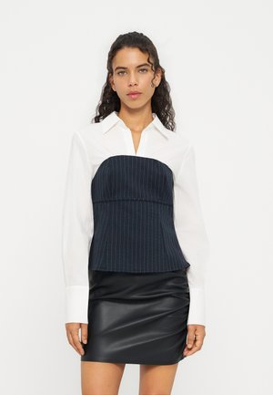Mujer con cabello rizado que lleva una camisa blanca con cuello, un corsé azul marino con rayas y una falda de cuero negra sobre un fondo blanco.