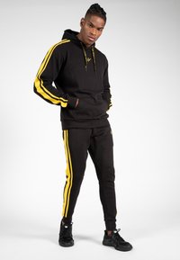 Zwarte hoodie en bijpassende broek met gele strepen aan de zijkanten. Het materiaal lijkt zacht en gestructureerd te zijn. Heeft een capuchon met trekkoord.