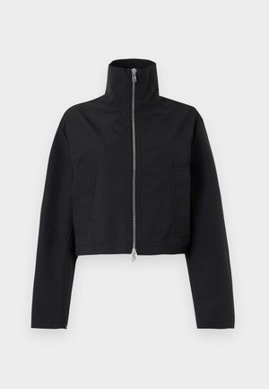 Kvinde står iført en sort cropped lynlåstrøje, hvid skjorte, blå viddebukser og sorte spidse højhælede sko på en ensfarvet baggrund.