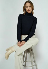 Navy-blau gerippter Rollkragenpullover mit langen Ärmeln, kombiniert mit cremefarbenen weit geschnittenen Hosen und beigen Knöchelstiefeln, sitzt auf einem Hocker.