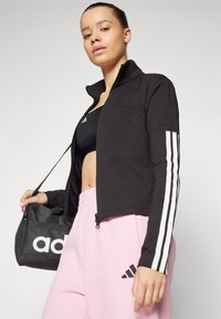 Črna atletska jakna s belimi črtami na rokavih, oblečena čez črn športni modrček, v kombinaciji z rožnatimi trenirkami in črno torbo.