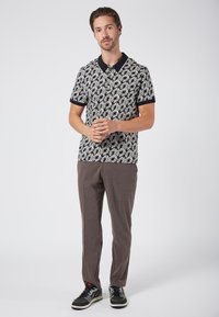 Kortärmad pikétröja med mönster av svarta och vita löv, svart krage, kombinerad med bruna texturerade byxor och svarta sneakers.