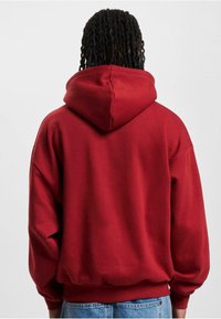 Rød bomullslue med romslig hette, ribbestrikkede mansjetter og nederkant, og senkede skuldrer, kombinert med blå denim bukser. Vist fra bak.