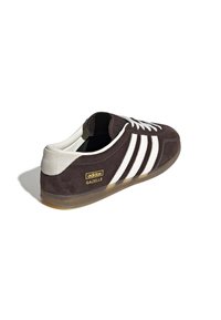 Καφέ σουέντ αθλητικό παπούτσι Adidas Gazelle με λευκές ρίγες, ανοιχτό γκρι τακούνι και σόλα καουτσούκ· διαθέτει λογότυπο σε χρυσό χρώμα.