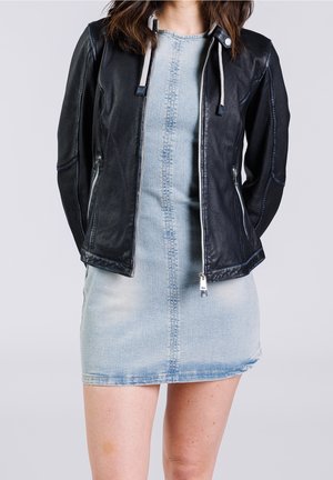 Femme portant une veste en cuir noire à fermeture éclair par-dessus une mini-robe en jean bleu clair délavé, debout avec les mains derrière le dos.