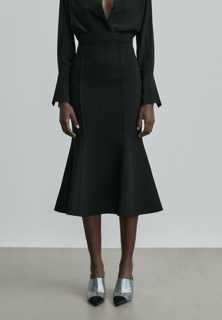 Femme portant une robe noire à manches longues avec une taille cintrée et un ourlet évasé, associée à des chaussures pointues argentées et noires.
