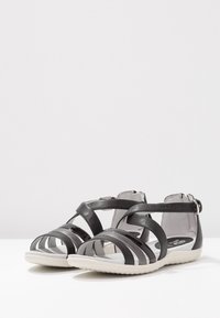 Mustat remmillä varustetut naisten sandaalit, joissa on valkoiset pohjat ja nilkkaremmit, esillä valkoisella pinnalla vaaleanharmaata taustaa vasten.