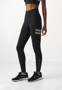 Tamno visoko pasu leggins v raztegljivem materialu, ki imajo stransko žep z vodoravnimi črtami in odbojnimi detajli pri gležnju.