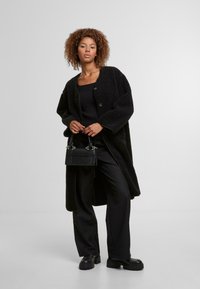 Schwarzer Teddy-Mantel mit großen vorderen Knöpfen, kombiniert mit einem schwarzen gerippten Oberteil und einer Hose. Hält eine kleine schwarze Tasche mit silbernen Akzenten.