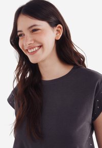 Kulkulgrå t-shirt med rund halsudskæring og korte ærmer, der har metalliske nitter på skuldrene. Glat bomuldsstruktur.