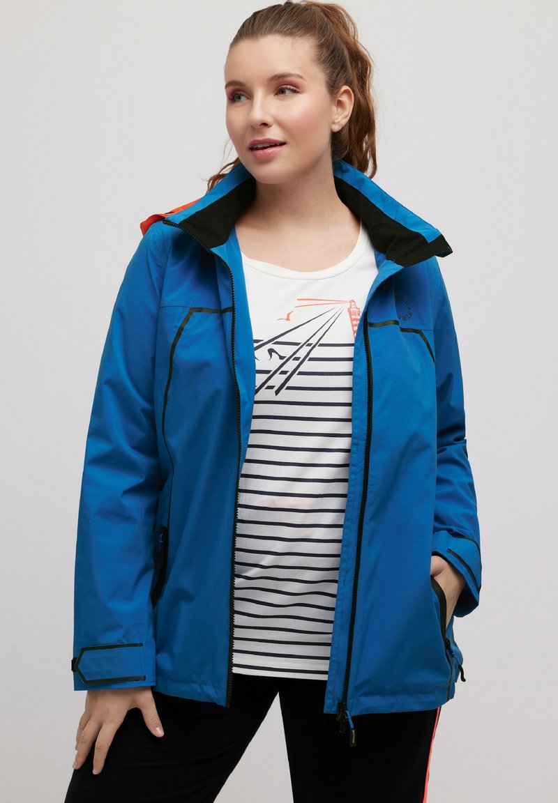 Blauwe waterdichte jas met een zwarte kraag, voorzien van ritszakken en contrasterende oranje accenten. Draag het over een gestreept shirt.