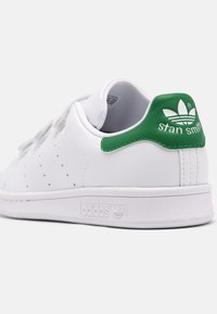 Baskets en cuir blanc avec des accents verts au niveau du talon, côtés perforés, semelle blanche plate, et logo Adidas embossé sur le côté.