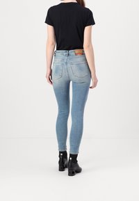 Persona vestida con jeans ajustados de color azul claro, camiseta negra de manga corta y botas de tacón negras hasta el tobillo, de pie y mirando hacia atrás sobre un fondo blanco.