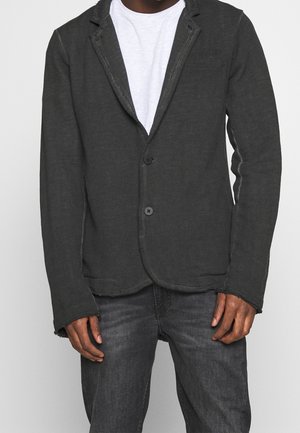 Man met een donkergrijze casual blazer over een wit T-shirt en donkergrijze jeans, halverwege de dij afgeknipt, staand tegen een effen achtergrond.