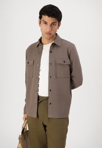 Camisa castanha de botão com dois bolsos no peito, botões pretos e uma textura suave, combinada com calças verde-oliva e uma mala bege.