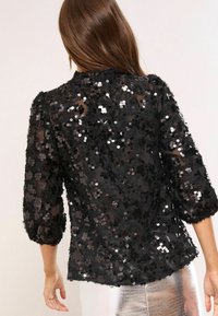 Must mustlane blouse, millel kaunistatud sädelevate detailide ja kolmeveerandvarrukatega. Seljaosa toob esile ümmarguse kaelaaugu.