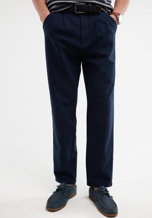 Pantalons bleu marine avec des plis à l'avant, des poches latérales et un ourlet à revers, associés à des chaussures bateau bleu foncé. Texture de tissu unie. Ceinturé.