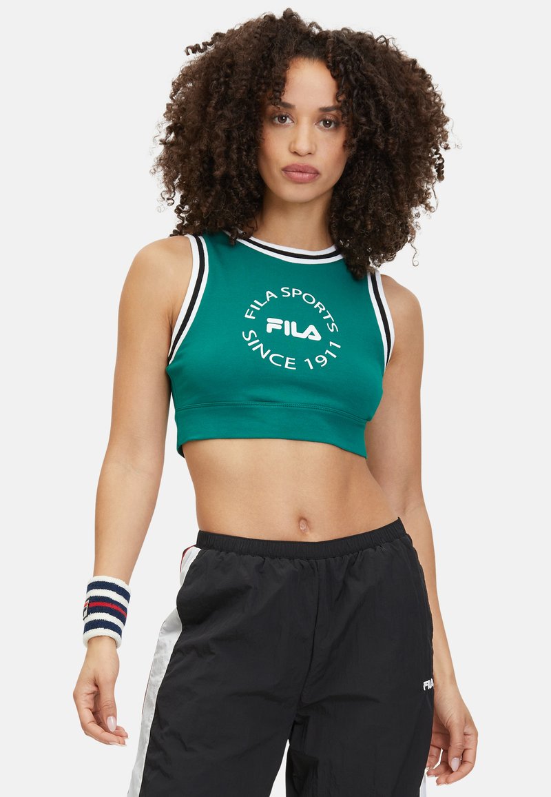 Mujer con cabello rizado, vistiendo un top deportivo corto verde de FILA, pantalones deportivos negros y blancos, y una banda de muñeca a rayas, de pie con una mano hacia abajo.