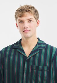 Námořnická modrá pyžamová košile se zelenými a červenými vertikálními pruhy, měkká tkanina, límec s výstřihem do V, přední kapsa a zapínání na knoflíky.