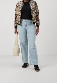 Bomberjacke mit Leopardenmuster, schwarzes geripptes Oberteil, hellblaue weite Jeans und schwarze Sneakers. Hält eine cremefarbene gepolsterte Schultertasche.