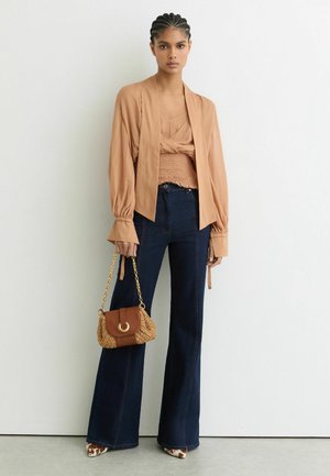 Kvinde med flettet hår iført en beige bluse, mørke vidde jeans, mønstrede hæle og holder en tan vævet håndtaske med kæderem.