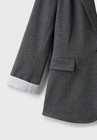 Manche de blazer gris foncé texturé avec un poignet de chemise blanc rayé visible au poignet et une poche à rabat sur le côté.