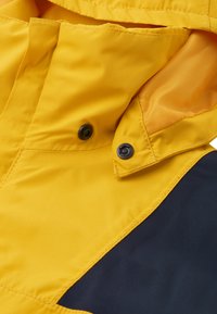Veste imperméable jaune avec une section contrastante bleu marine, dotée d'une capuche, de boutons-pression et d'une texture lisse. Détails cousus visibles.
