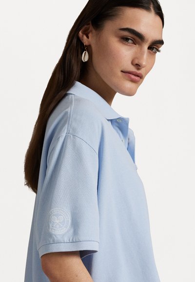 Šviesiai mėlyna polo marškinėliai su trumpomis rankovėmis, pagaminti iš tekstūruotos medžiagos, turi logotipą ant rankovės ir klasikines apykaklę.