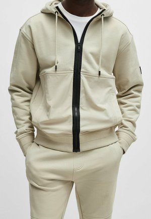 Man som bär en beige zip-up huvtröja med svarta dragkedjor och matchande beiga byxor, med händerna i fickorna och en vit skjorta under.