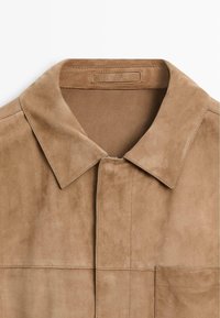 Hellbrauner Wildlederjackenkragen mit sichtbaren Nähten und einer rechteckigen Brusttasche auf der rechten Seite.