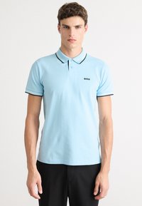 Polo bleu clair avec un logo noir, présentant un col classique, une patte de boutonnage à deux boutons et des finitions contrastantes sur les manches.