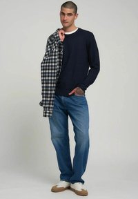 Maglione blu navy, camicia a quadri e jeans blu. La camicia presenta quadretti bianchi e neri, mentre le scarpe sono bianche con dettagli marroni.