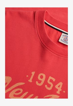 T-shirt rouge à col rond avec le chiffre "1954" beige et du texte partiellement en cursive, avec une étiquette Superdry Outfitter à l'intérieur du col.