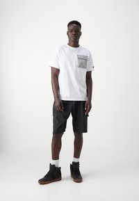 Hvid bomulds t-shirt med grå lomme detalje, parret med sorte cargoshorts og sorte høj-top sneakers. Enkel og funktionel design.