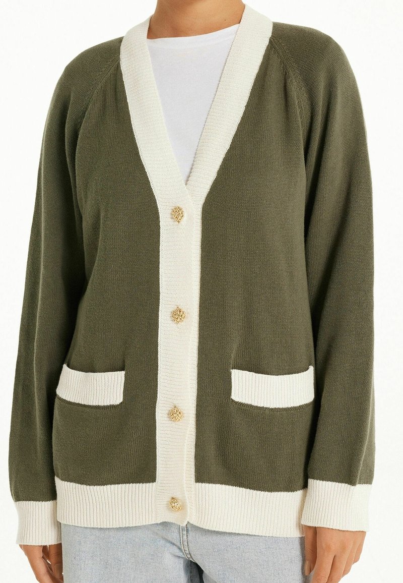 Cardigan vert olive avec bordure blanche, boutons dorés texturés, deux poches avant, porté sur un haut blanc et un jean bleu clair.